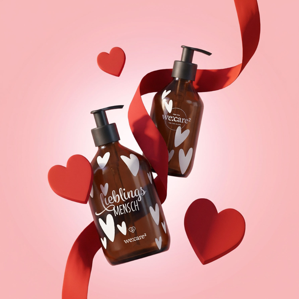 Braune Seifenspender von we:care² mit weißen Herz‑Stickern, umgeben von roten 3D‑Herzen und einem geschwungenen roten Band vor einem rosa Hintergrund.